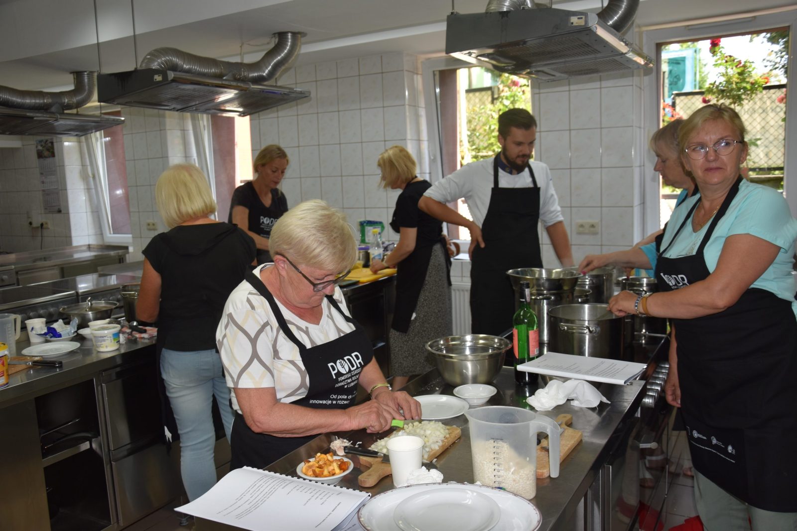 Grupa uczestników warsztatów kulinarnych, zarówno kobiety, jak i prowadzący w fartuchach, przygotowują dania w kuchni. Kobieta z przodu kroi cebulę, a w tle pozostałe osoby są zaangażowane w różne czynności kulinarne. Widać świeże składniki oraz garnki i naczynia używane podczas warsztatów.